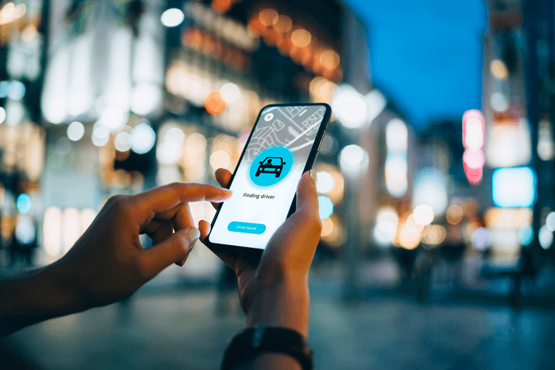 Person stands near a busy street at dusk while using a smartphone to request a car service.